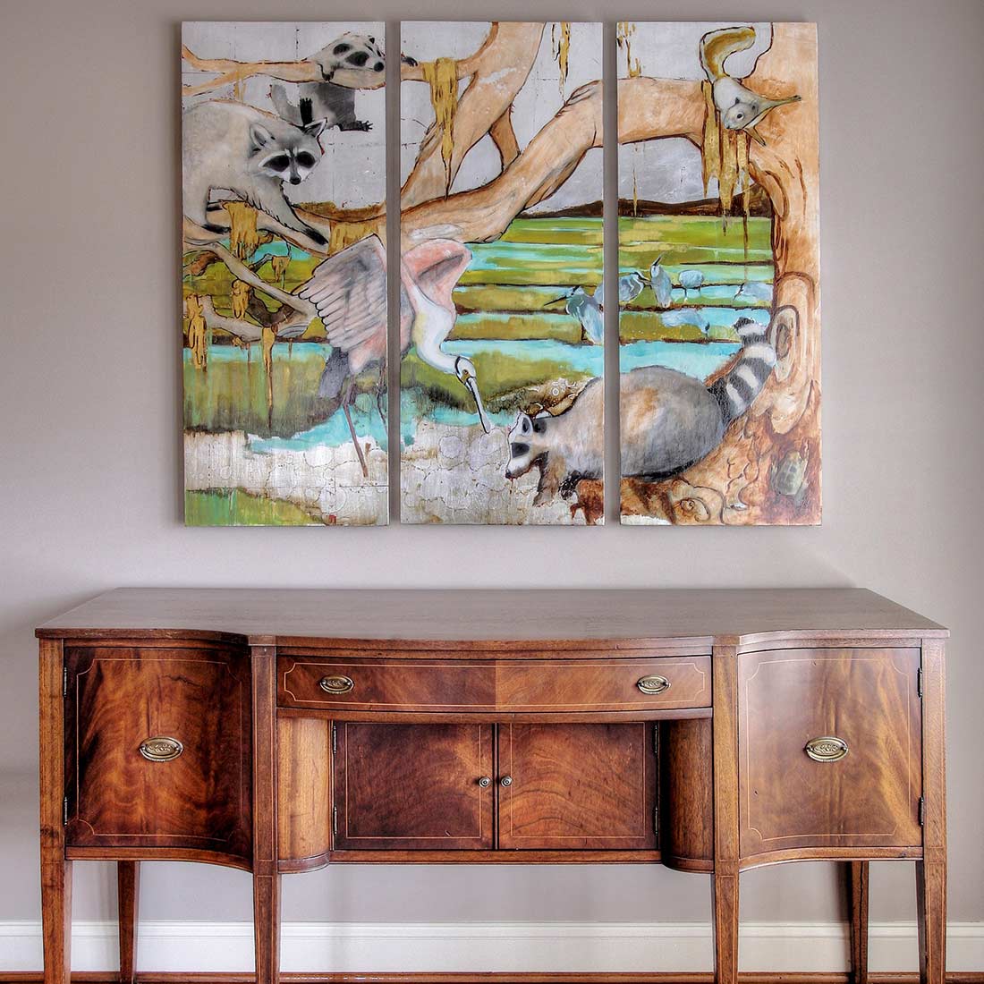 Elegant wooden sideboard with intricate veneer patterns, featuring brass handles, styled with a triptych painting above showcasing raccoons and a heron in a natural setting.